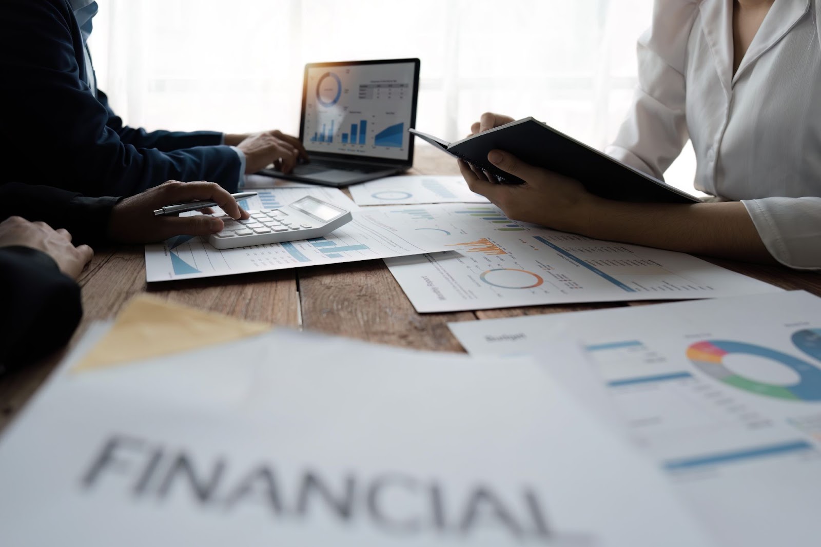 A hand holding a silver pen points to a pie chart, analyzing a business's financial report. The table also features additional reports, a coffee cup, a calculator, and a laptop.