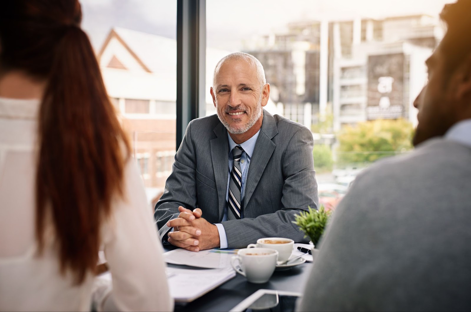 A male professional consultant in a gray business suit smiles warmly at a couple whose backs are to the camera as they discuss and envision future business success together.