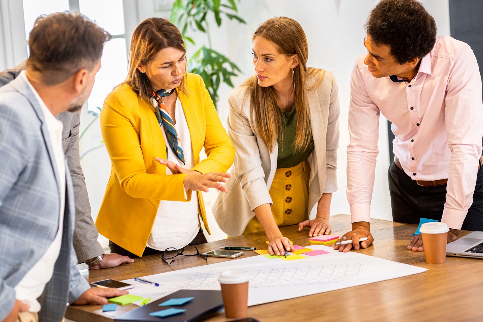 A female management consultant, elegantly attired in a yellow blazer with a white inner layer and a blue scarf, discusses strategies with three business staff members.