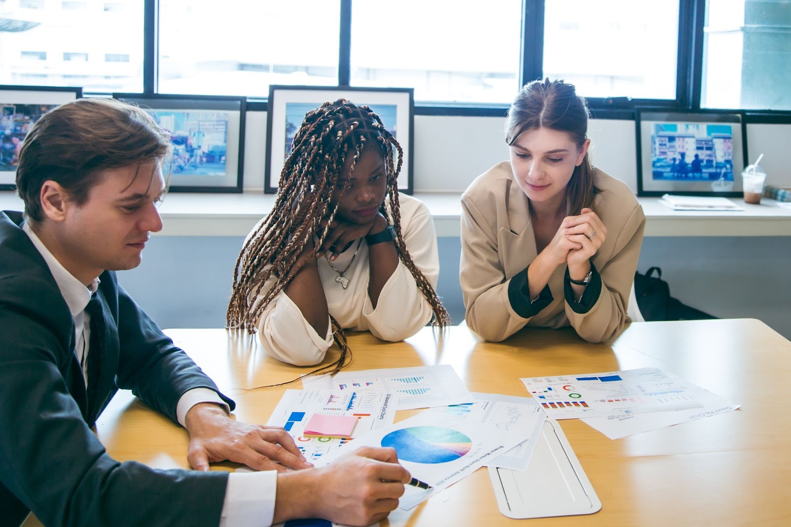 Three professional consultants engaged in a strategy discussion, examining statistical documents at a table, with picture frames visible on shelves in the background.