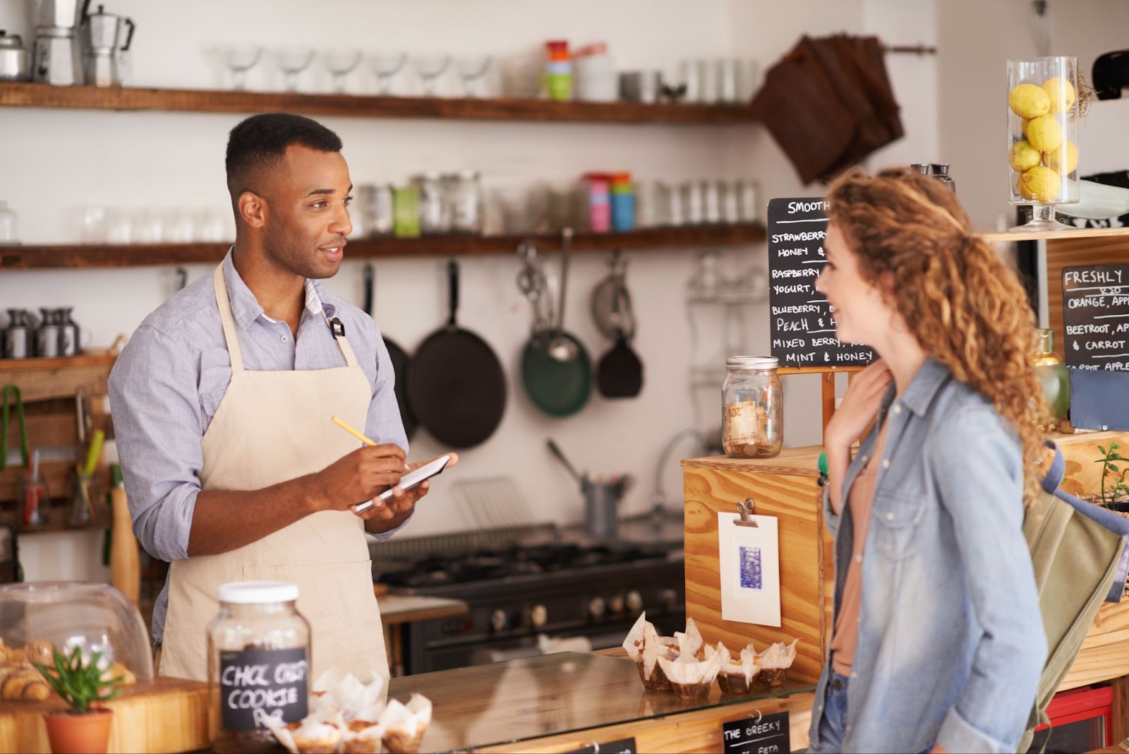 A male coffee shop employee attentively takes orders from a female customer at the counter, with café essentials arranged in the background, contributing to the cozy ambiance of the café.