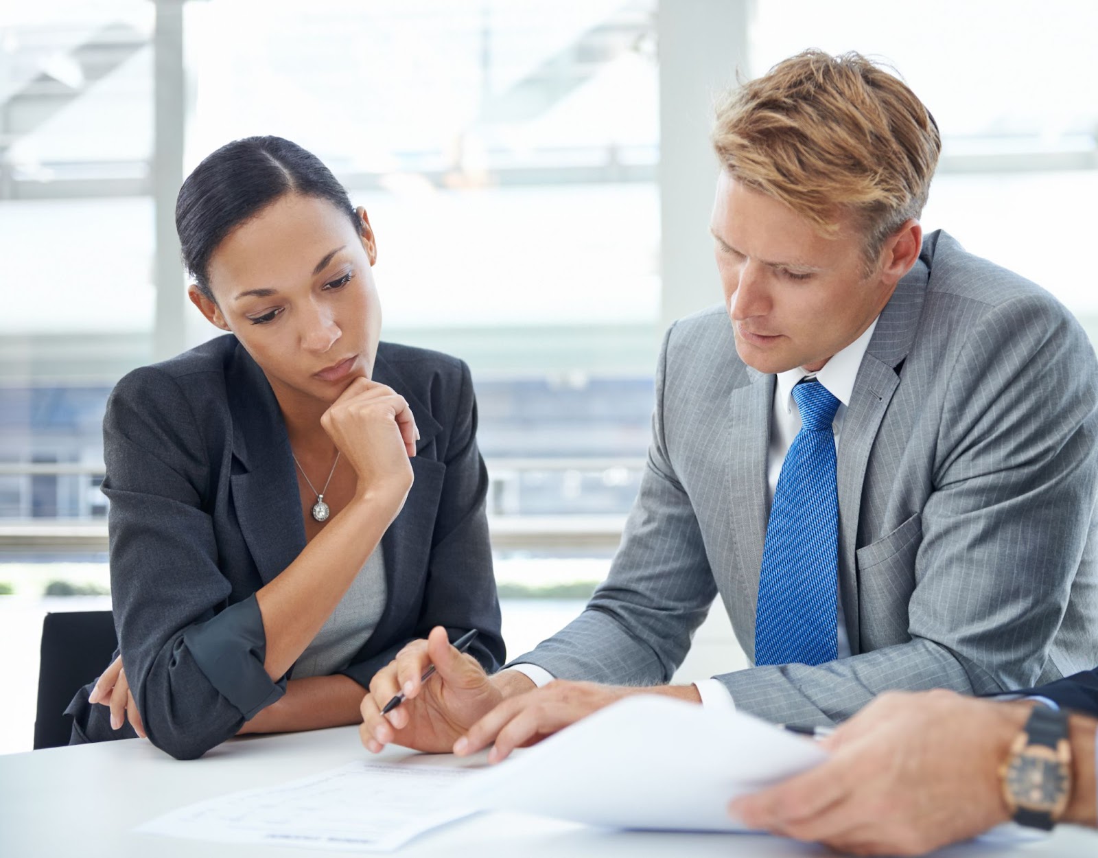 A Caucasian business consultant in a light gray suit and blue necktie reviews assets and liabilities with a female executive in a black suit. A male hand wearing a watch is also visible.