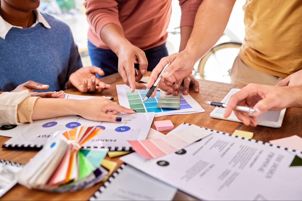 Several hands of a diverse creative team collaborating on the product development process, with documents, swatches, and strategy materials visible as they work on a project together.