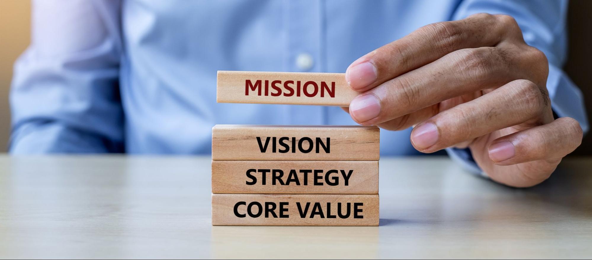 A businessman's hand in a light blue long-sleeved polo arranging wooden blocks labeled mission, vision, strategy, and core values, representing business goals alignment.