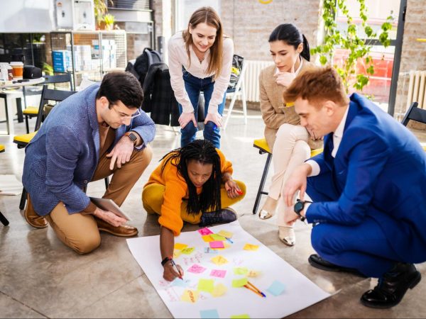 A business team uses sticky notes on a large white cartolina on the floor to brainstorm and develop a profitable business idea.