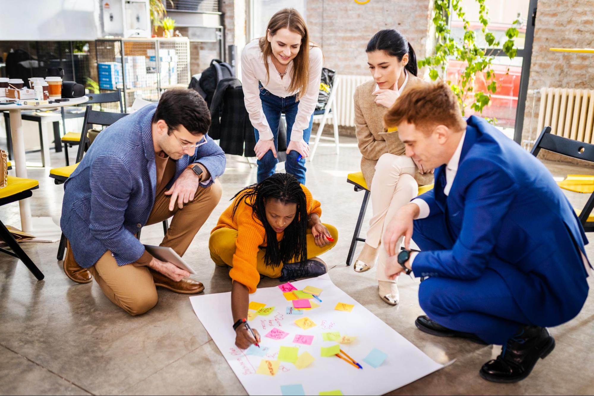 A business team uses sticky notes on a large white cartolina on the floor to brainstorm and develop a profitable business idea.