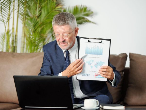 A male business advisor wearing a suit discusses marketing strategy via video call, showing a chart on a clipboard.