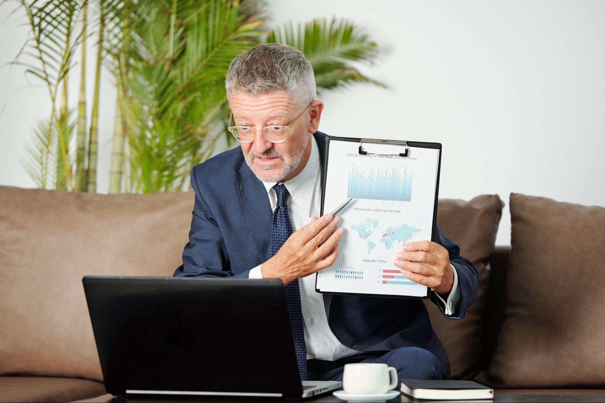 A male business advisor wearing a suit discusses marketing strategy via video call, showing a chart on a clipboard.