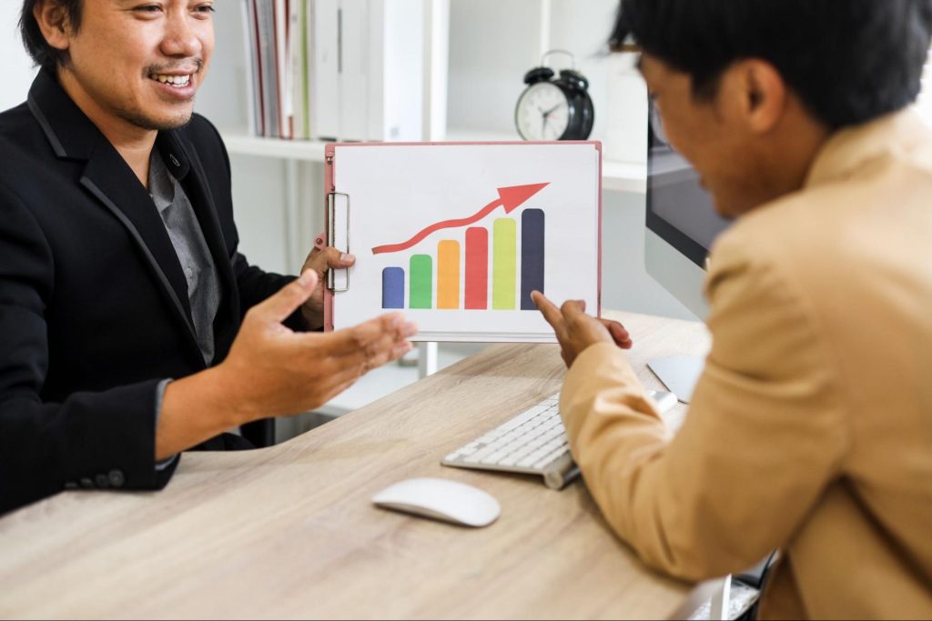 A businessman discusses sales growth with his colleague, holding a colorful graph featuring an upward arrow above.